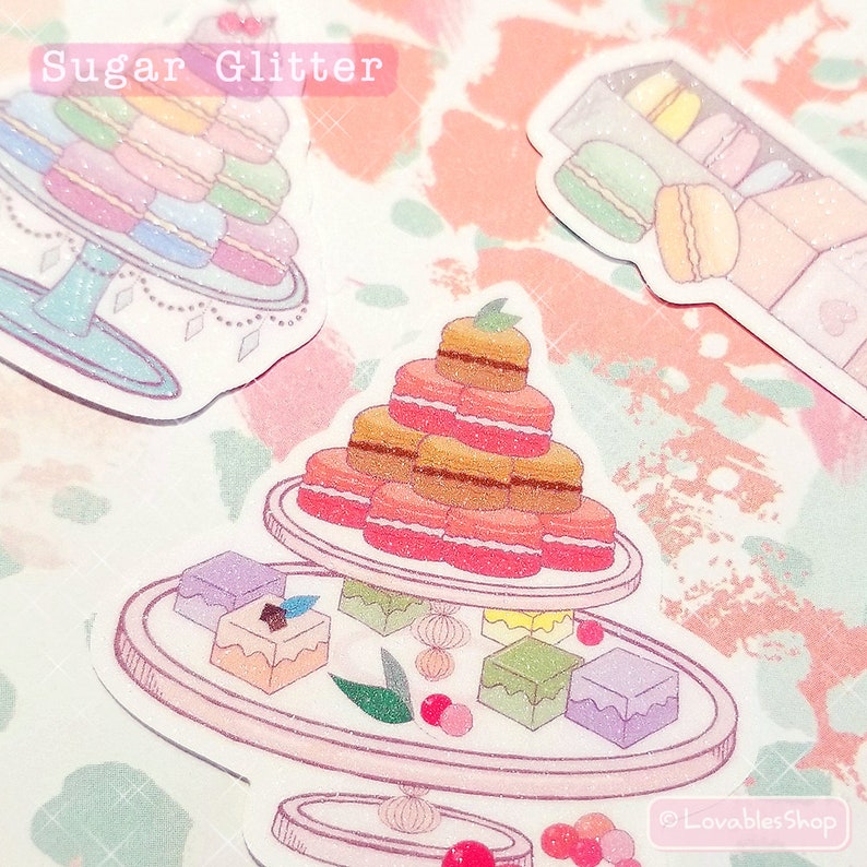 May include: A set of three pink and white stickers featuring a tower of macarons, a tiered tray of macarons and other pastries, and a shelf of macarons. The stickers have a sparkly glitter finish and the text "Sugar Glitter" is at the top of the image.