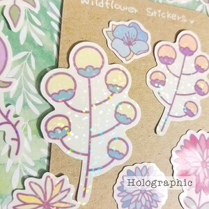 May include: A sheet of holographic stickers featuring various flower designs. The stickers are on a brown background with the text "Wildflower Stickers" and "Holographic" printed on the sheet.