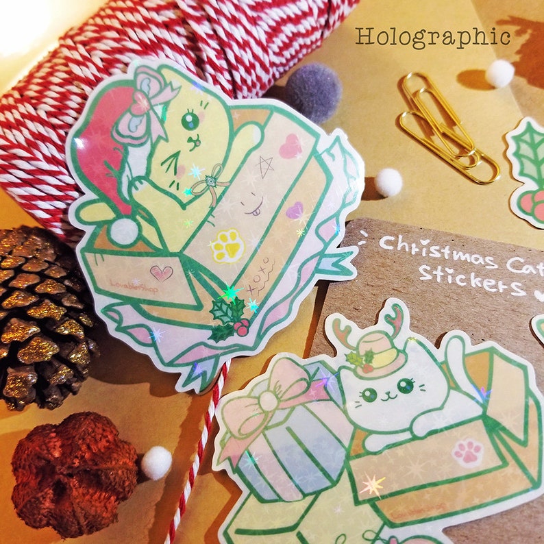 May include: Two holographic Christmas cat stickers. One sticker features a yellow cat wearing a Santa hat in a box with a red heart and holly. The other sticker features a white cat wearing a hat with antlers in a box with a red bow and holly. The text "Christmas Cat Stickers" is written on the brown paper background.