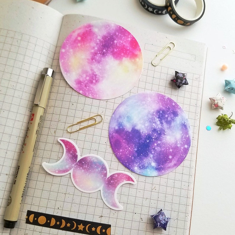 May include: Three galaxy-themed stickers, one with a triple moon symbol and two with full moons. The moons are pink, purple, and blue with white and yellow accents.