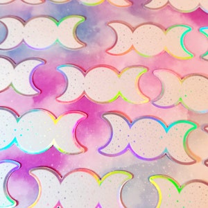 May include: A sheet of iridescent holographic stickers featuring the triple moon symbol. The stickers are white with a glittery finish and have a rainbow iridescent border.