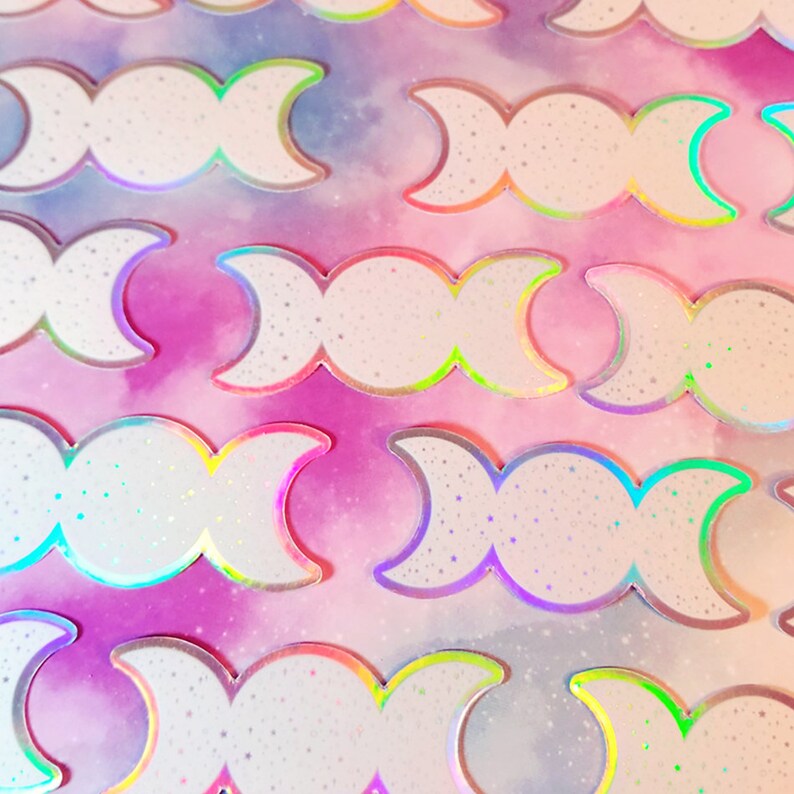 May include: A sheet of iridescent holographic stickers featuring the triple moon symbol. The stickers are white with a glittery finish and have a rainbow iridescent border.