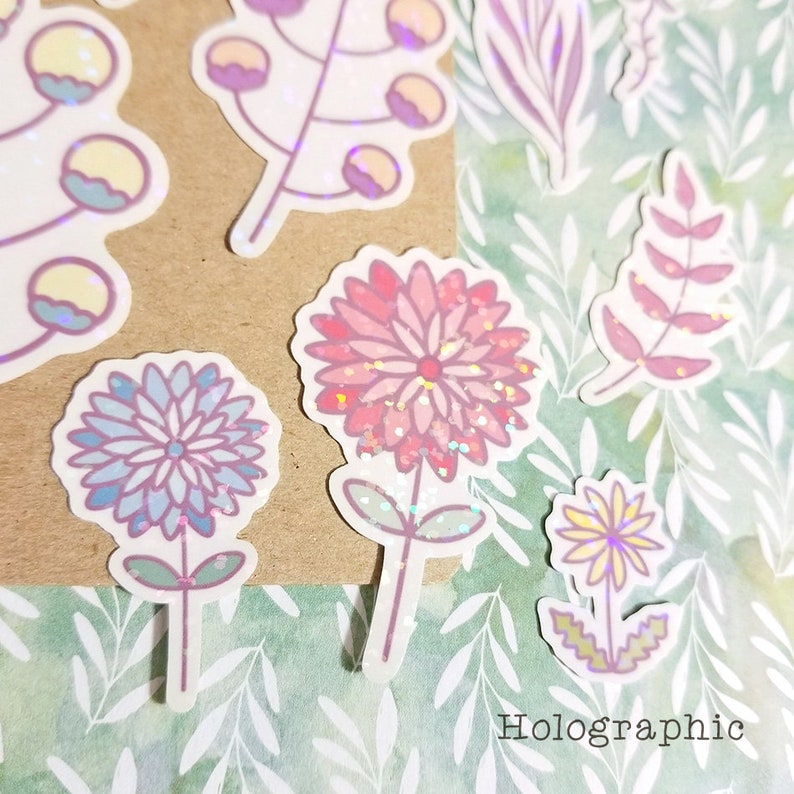 May include: A set of holographic floral stickers featuring pink, blue, and yellow flowers with glitter accents. The stickers are on a green and white background with the word "Holographic" printed at the bottom.
