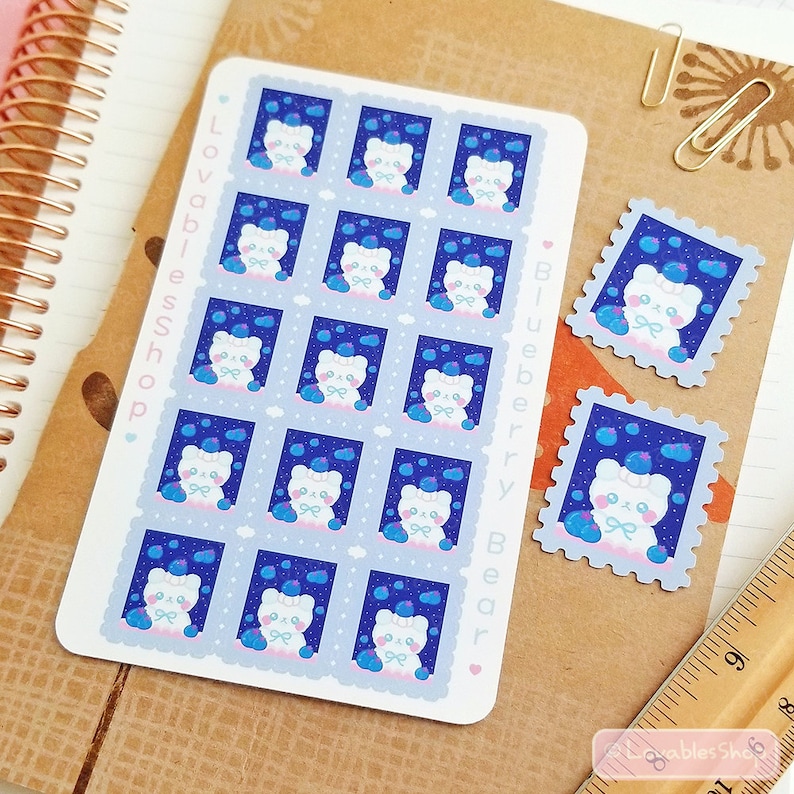 May include: A sheet of 12 stickers featuring a white bear with blue eyes and a blue bow, surrounded by blue blueberries on a blue background. The stickers are in the shape of a postage stamp with a white border. The sheet is labeled "Blueberry Bear" and "LovablesShop."