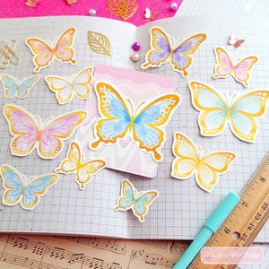 May include: A sheet of colorful butterfly stickers with gold accents. The stickers are arranged on a gridded notebook page with a ruler and a pen. The text "Butterfly Stickers" is visible on one of the stickers.