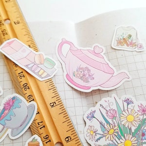 May include: A set of pastel pink and green stickers featuring a teapot, a tea bag, macarons, a cup of tea, and a bouquet of flowers. The stickers are on a white gridded background.