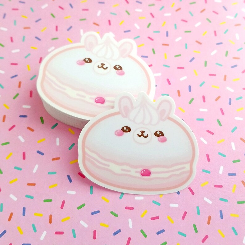May include: Two pink and white macaron stickers with cute cartoon llama faces. The macarons have pink frosting and a red cherry on top. The stickers are on a pink background with colorful sprinkles.