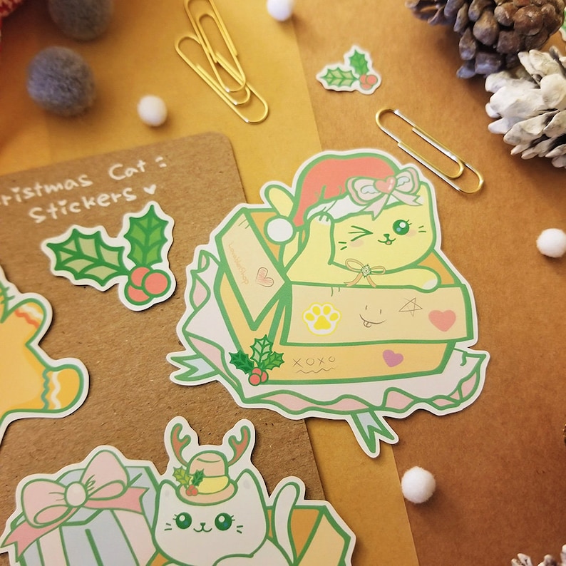 May include: A set of Christmas cat stickers featuring a yellow cat wearing a Santa hat in a box, a white cat wearing a reindeer hat, and a green holly sprig. The stickers are on a brown background.