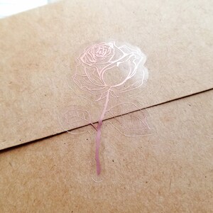 May include: A rose sticker with a rose and leaves in a rose gold color on a clear background.