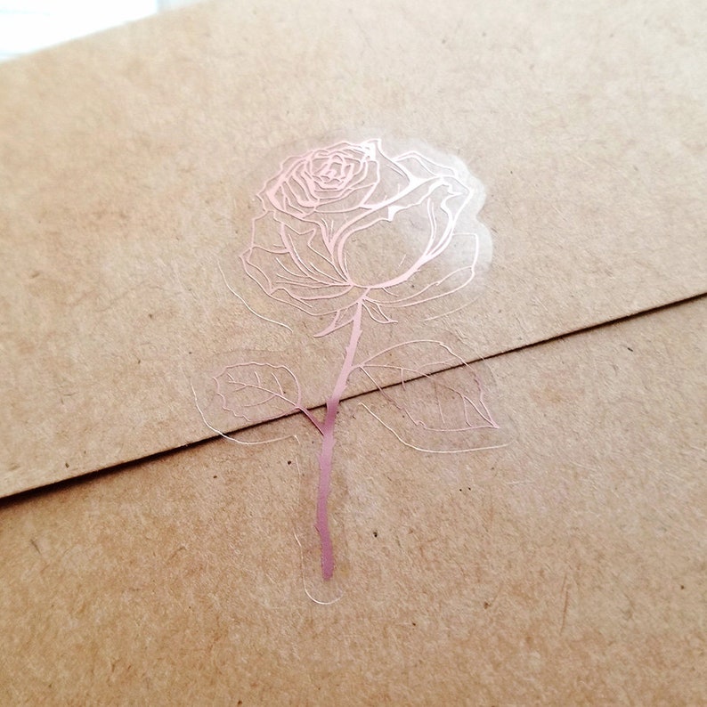 May include: A rose sticker with a rose and leaves in a rose gold color on a clear background.