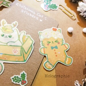 May include: Two holographic Christmas cat stickers. One sticker features a white cat wearing a Santa hat and sitting in a box. The other sticker features a gingerbread cat wearing a blue bow tie. The text "Christmas Cat Stickers" and "Holographic" are visible on the brown background.