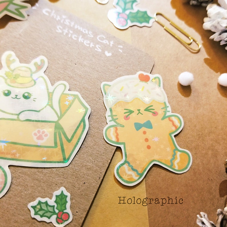 May include: Two holographic Christmas cat stickers. One sticker features a white cat wearing a Santa hat and sitting in a box. The other sticker features a gingerbread cat wearing a blue bow tie. The text "Christmas Cat Stickers" and "Holographic" are visible on the brown background.