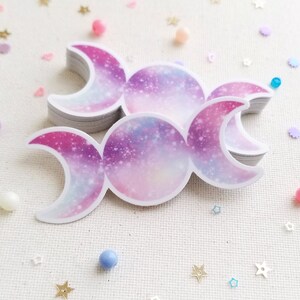 May include: A stack of triple moon stickers with a galaxy design in shades of pink, purple, and white.