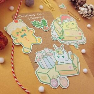 May include: A set of four Christmas cat stickers featuring a gingerbread cat, a cat in a box with a Santa hat, a cat in a box with reindeer antlers, and a cat in a box with a gift. The stickers are on a brown background.
