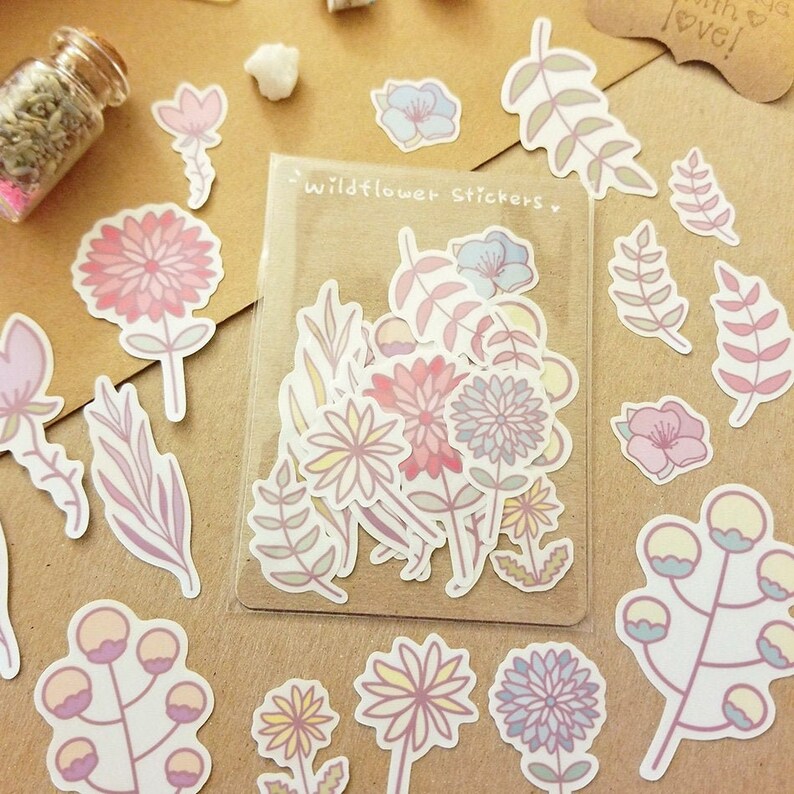 May include: A sheet of stickers featuring various pastel-colored wildflowers and foliage. The stickers are packaged in a clear plastic sleeve with the text "Wildflower Stickers" printed on it.