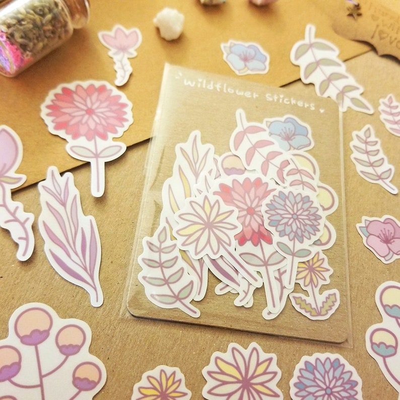 May include: A sheet of stickers featuring a variety of pastel-colored wildflowers and leaves. The stickers are packaged in a clear plastic sleeve with the text "Wildflower Stickers" printed on the front.
