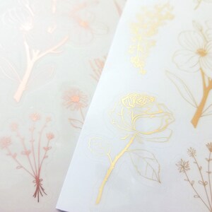 May include: Two sheets of clear sticker paper with gold and rose gold floral designs. The designs include a rose, leaves, and various small flowers.