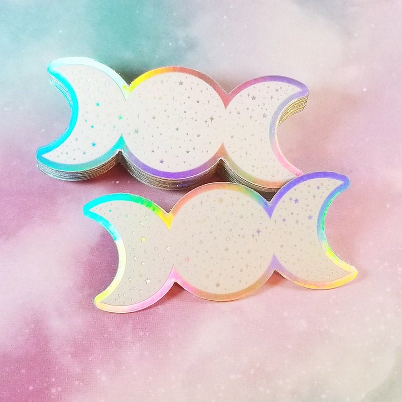 May include: A stack of iridescent holographic stickers featuring the triple moon symbol. The stickers are white with a glittery finish and have a rainbow iridescent border.