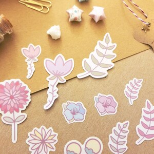May include: A set of pastel pink, purple, and blue floral stickers with green leaves. The stickers are on a brown paper background.