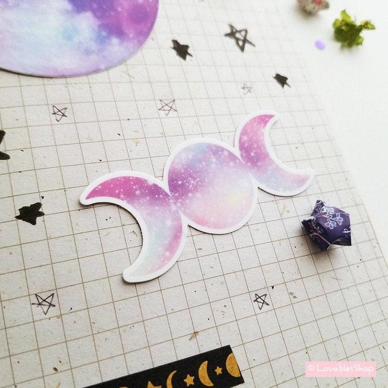 May include: A triple moon sticker with a galaxy print in shades of pink, purple, and blue. The sticker is on a gridded background.
