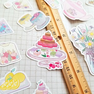 May include: A set of colorful stickers featuring various designs, including a stack of macarons, a bouquet of flowers, a teacup, and a lemon slice. The stickers are on a wooden ruler with inch markings.