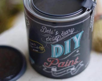 May include: A black cylindrical paint can with a metal rim and lid. The label features white, teal, and pink text that reads Debi's design diary DIY Paint. A small black sticker is attached to the top rim, and a round brush is visible nearby.