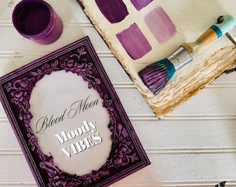May include: A purple paint jar, a paintbrush with purple-stained bristles, and a rectangular frame with floral relief details. The frame displays the text Blood Moon Moody VIBES. An open book shows four distinct shades of purple paint swatches.