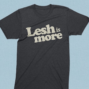 May include: A dark heather grey t-shirt with the text "Lesh is more" printed in white.