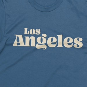 May include: A blue t-shirt with the text "Los Angeles" in a retro, cream-colored font. The shirt has a crew neck and is made of a soft material. The design is centered on the chest.