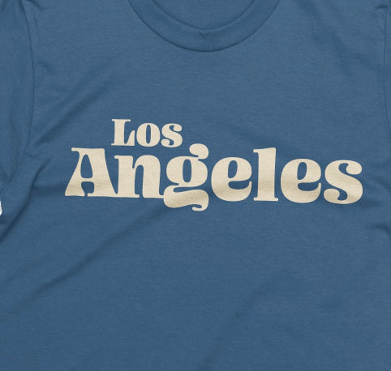 May include: A blue t-shirt with the text "Los Angeles" in a retro, cream-colored font. The shirt has a crew neck and is made of a soft material. The design is centered on the chest.