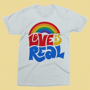 May include: A white t-shirt with a rainbow graphic and the words "LOVE IS REAL" in red, yellow, and blue. The design features a rainbow arching over the text, creating a vibrant and colorful statement. The shirt is a classic crew neck style.