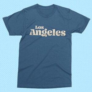 May include: A blue t-shirt with the text "Los Angeles" in a retro font. The text is in a light beige color and is centered on the front of the shirt. The shirt has short sleeves and a crew neck.