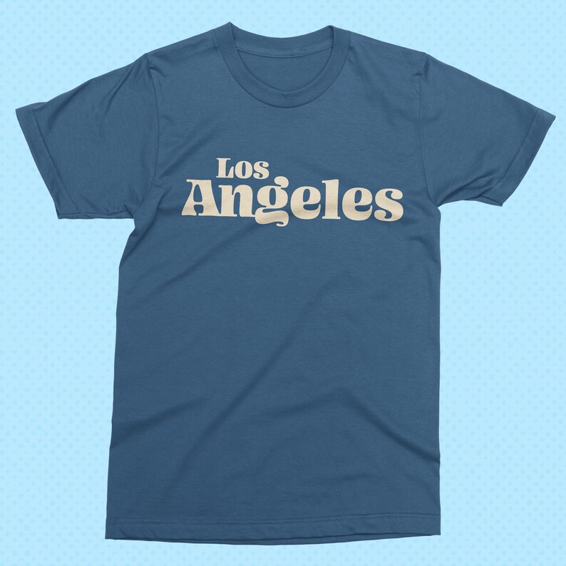 May include: A blue t-shirt with the text "Los Angeles" in a retro font. The text is in a light beige color and is centered on the front of the shirt. The shirt has short sleeves and a crew neck.