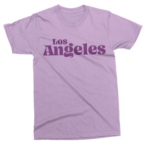 May include: A lilac-colored t-shirt with the text "Los Angeles" printed in a darker purple font across the chest. The text has a retro, rounded font style. The shirt has short sleeves and a crew neck.