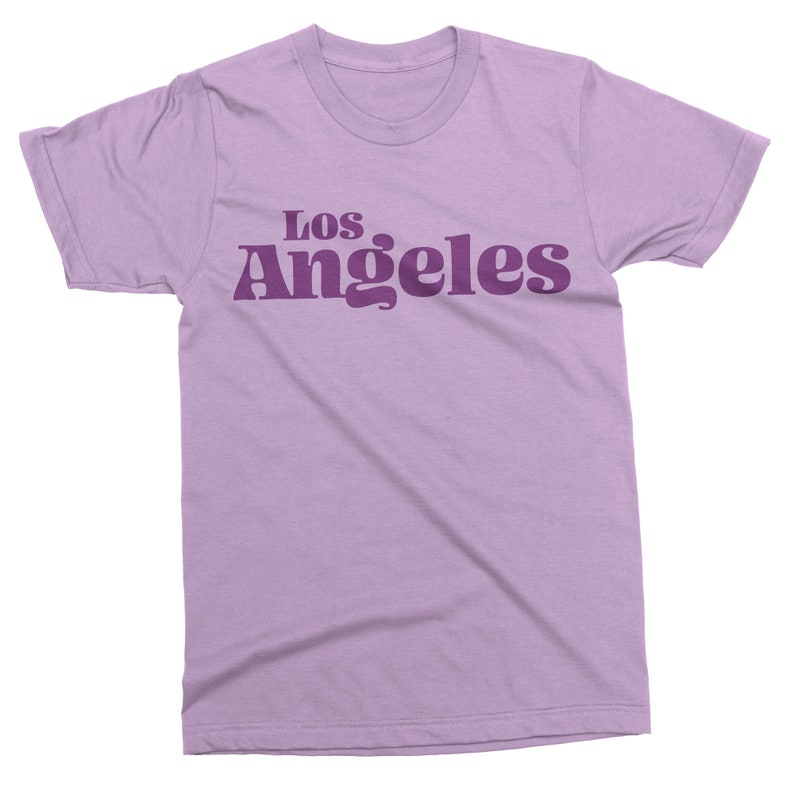 May include: A lilac-colored t-shirt with the text "Los Angeles" printed in a darker purple font across the chest. The text has a retro, rounded font style. The shirt has short sleeves and a crew neck.