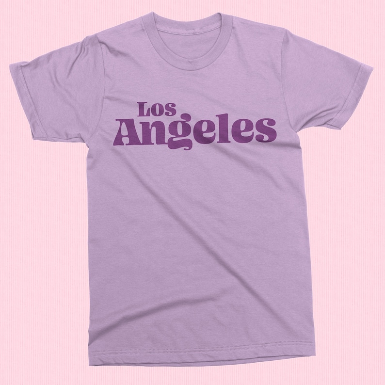 May include: A light purple t-shirt with the text "Los Angeles" in a darker purple, retro-style font. The shirt has a crew neck and short sleeves. The background is a light pink.
