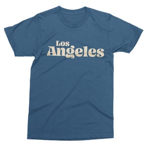 May include: A blue t-shirt with the text "Los Angeles" in a retro font. The text is in a light beige color and is centered on the front of the shirt. The shirt has short sleeves and a crew neck.