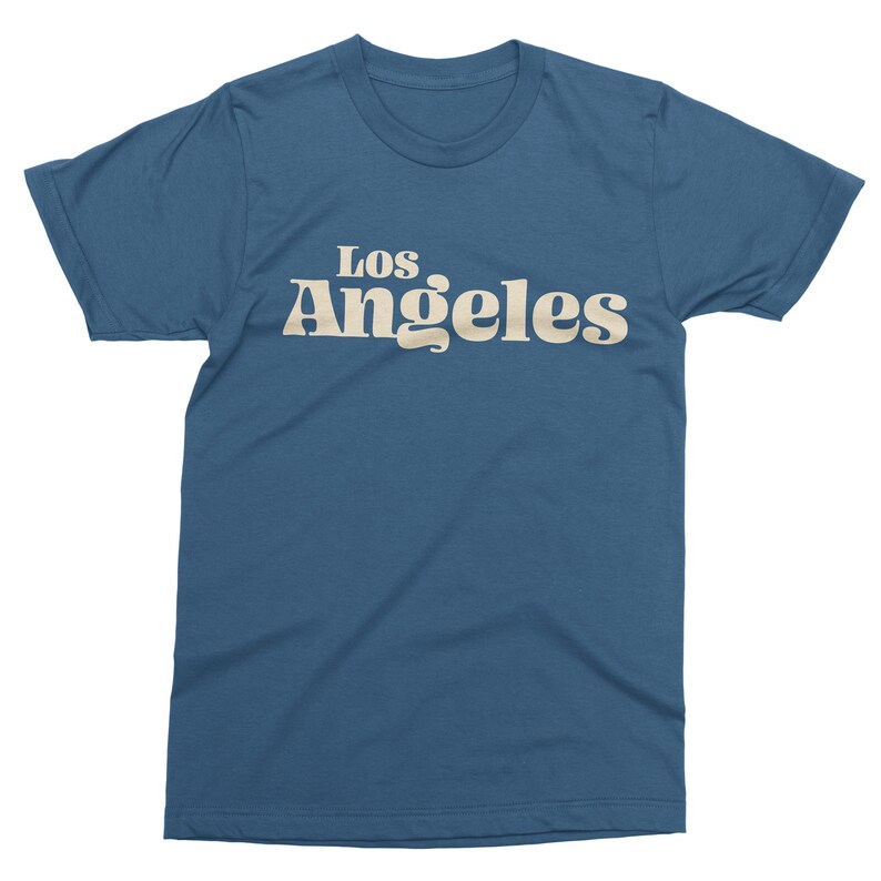 May include: A blue t-shirt with the text "Los Angeles" in a retro font. The text is in a light beige color and is centered on the front of the shirt. The shirt has short sleeves and a crew neck.