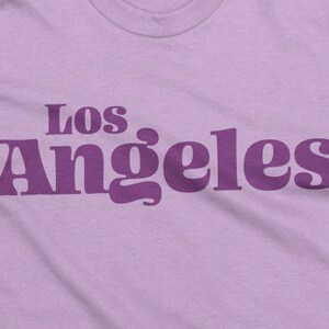 May include: A light purple t-shirt with the text "Los Angeles" printed in a darker purple, retro-style font. The shirt has a crew neck and is made of a soft fabric.