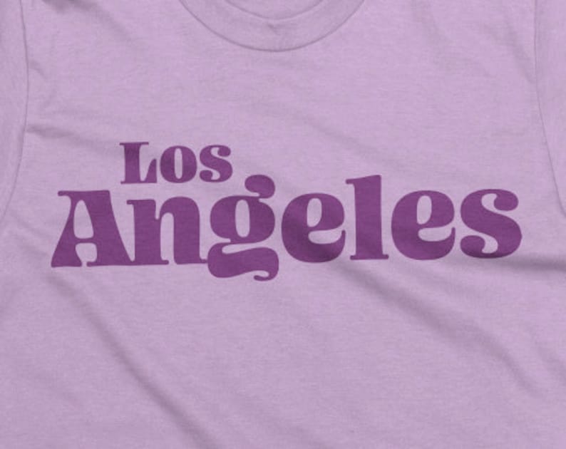 May include: A light purple t-shirt with the text "Los Angeles" printed in a darker purple, retro-style font. The shirt has a crew neck and is made of a soft fabric.