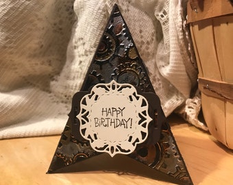 May include: A brown and black triangular card with a white lacy circle in the center. The circle has the text "Happy Birthday!" inside. The card has a steampunk design with gears and metallic accents.