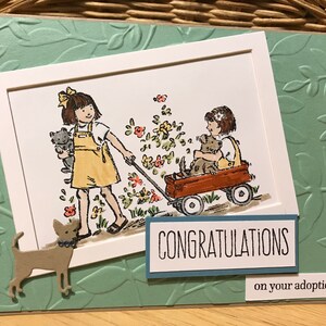 May include: A greeting card featuring a light green embossed leaf background. A central illustration shows two children in yellow overalls, one pulling a red wagon with a dog inside, the other holding a cat. Text reads CONGRATULATIONS on your adoption.