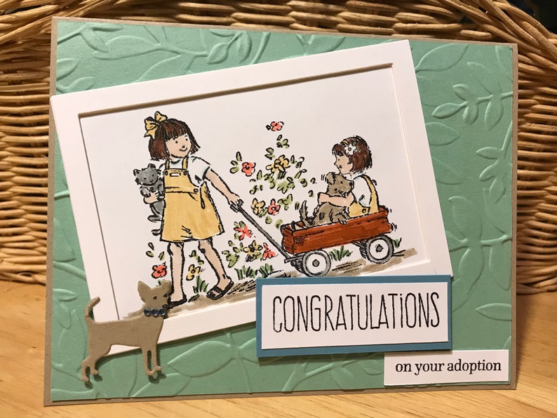 May include: A greeting card featuring a light green embossed leaf background. A central illustration shows two children in yellow overalls, one pulling a red wagon with a dog inside, the other holding a cat. Text reads CONGRATULATIONS on your adoption.