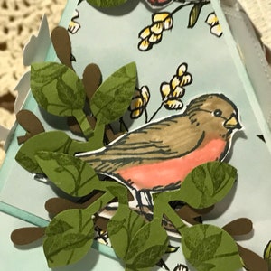 May include: A handmade papercraft gift box featuring a bird and branch design. The bird is brown and red, perched on a branch with green leaves. The box is light blue with additional paper cutouts of leaves and birds.