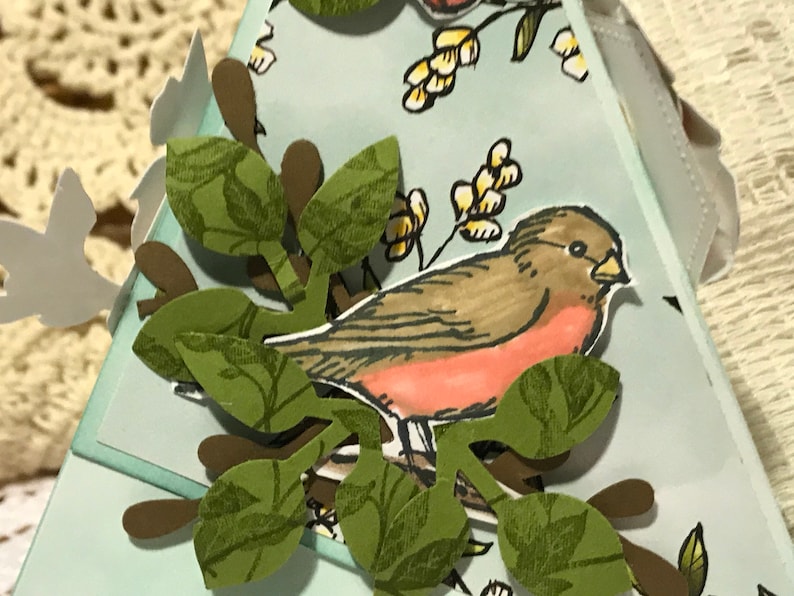 May include: A handmade papercraft gift box featuring a bird and branch design. The bird is brown and red, perched on a branch with green leaves. The box is light blue with additional paper cutouts of leaves and birds.