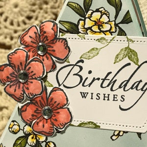 May include: A handmade birthday card with coral flower embellishments and sparkling rhinestones. The card features the words "Birthday Wishes" in elegant script on a white, stitched-edge label, with a floral background.