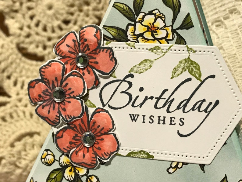 May include: A handmade birthday card with coral flower embellishments and sparkling rhinestones. The card features the words "Birthday Wishes" in elegant script on a white, stitched-edge label, with a floral background.