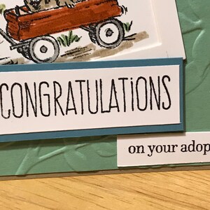 May include: A greeting card featuring a hand-drawn illustration of a brown wagon with a dog inside, set against a sage green embossed background. The card displays the text CONGRATULATIONS in black capital letters and on your adoption below.