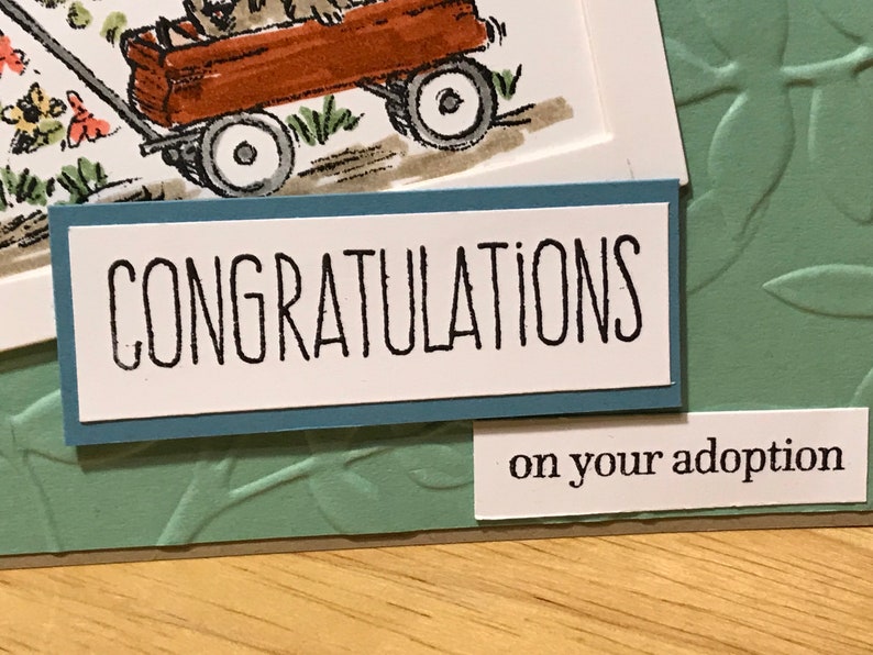 May include: A greeting card featuring a hand-drawn illustration of a brown wagon with a dog inside, set against a sage green embossed background. The card displays the text CONGRATULATIONS in black capital letters and on your adoption below.