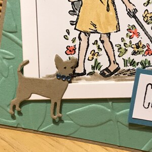 May include: A handmade greeting card featuring a die-cut tan dog with three small blue rhinestones on its collar. The card has a light green embossed leaf background and a white panel with a partial illustration of a person and the text CONG.